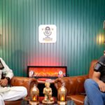 Two guests recording on a sofa with a decorative fireplace and green backdrop